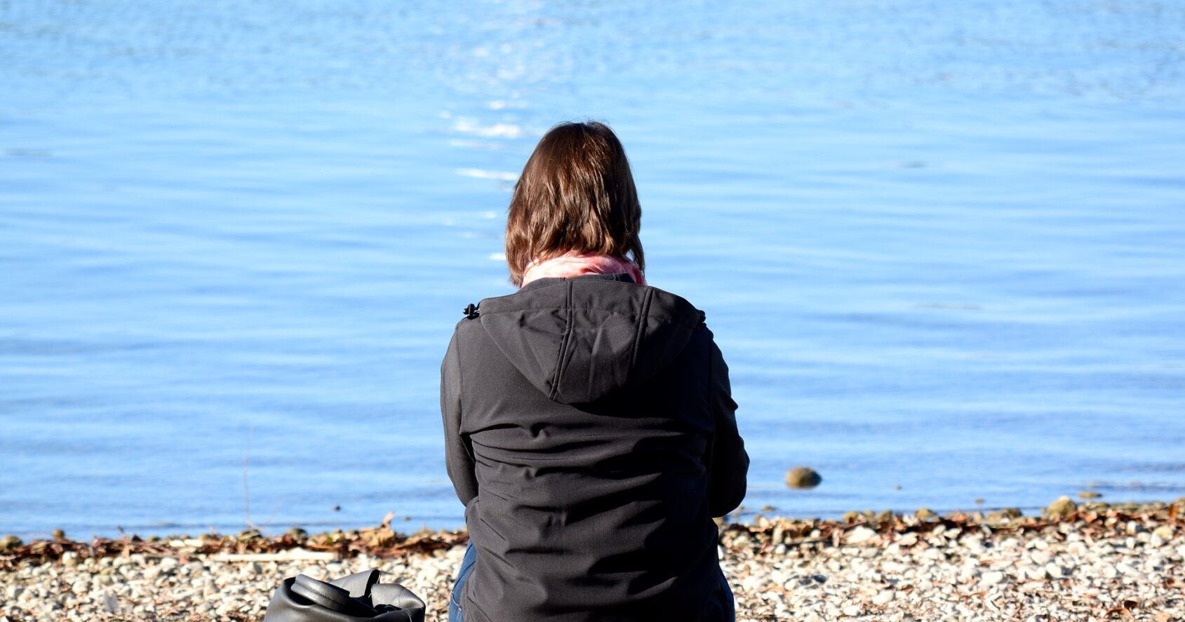 Järven rannalla selin kameraan kumarassa istuva henkilö