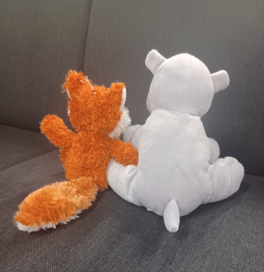 Kaksi pehmolelua istuu selin sohvalla toisiinsa tukeutuen.